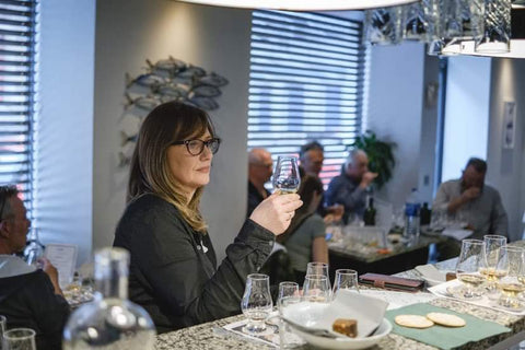 Mary Faller co owner of the Island Spirit Whisky Shop sitting at a whisky tasting holding a glass to eye height examining the contents.