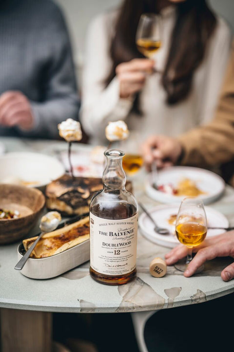 A bottle of The Balvenie Double Wood 12 Year Old whisky sits on a dining table surrounded by food and people holding whisky glasses in a softly blurred indoor setting.