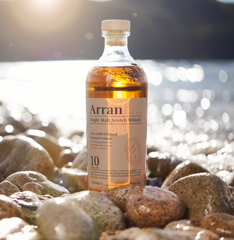 Bottle of Arran 10 year old single malt whisky sitting on a rocky shoreline with a bright sun and island in the background. Pale label with Arran branding. Whisky is amber / gold in colour. 