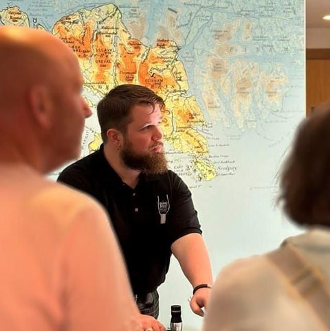 A picture of Conner Macaulay staff member of the island spirit whisky shop with a large map in the background and a couple of people blurred in the fore ground. 