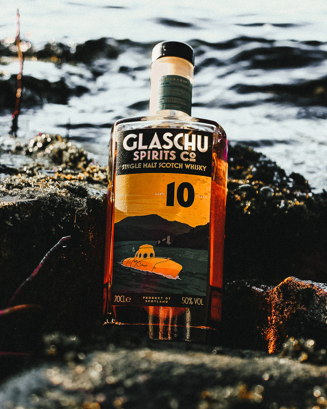 A bottle of Glaschu spirits co single malt whisky sitting on some rocks with the sea in the background. The label has the company branding and imagery showing a yellow submarine surfacing in a loch surrounded by mountain and a bright yellow sky.  