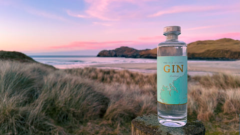 A bottle of the isle of lewis gin with a teal label sitting on a fence post over looking a beach with a pinky red sunset 