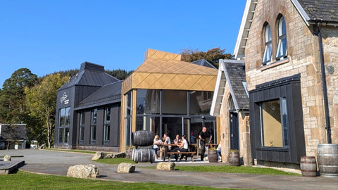 A photo of the front of the isle of Raasay distillery taken on a bright sunny day. Picnic bench with people sitting and enjoying a dram in the distant with casks out in front of the distillery. 