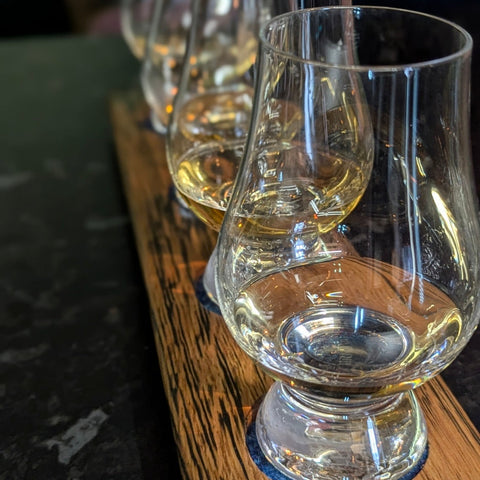 A tasting flight of 4 drams sitting on a whisky cask stave with slots made to hold the drams. Each slot has Harris tweed inserts for the glass to sit on. Bar scene in the background. 